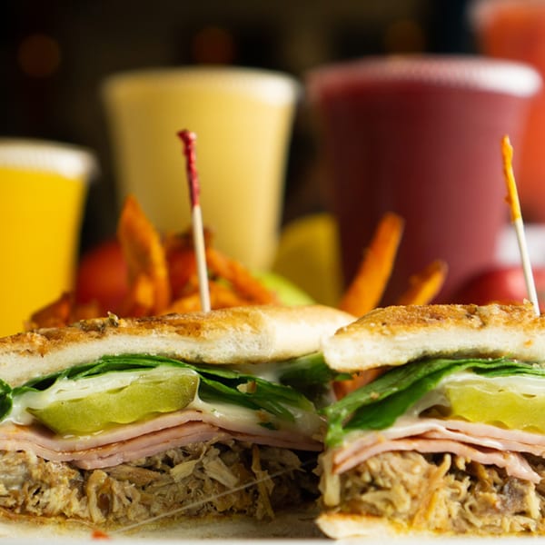 Close-up of a Cuban sandwich with pulled pork, ham, pickles, and lettuce, with drinks in the background.
