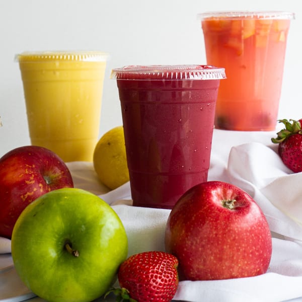 Three colorful Lincoln Grill Cafe smoothies with fresh apples, lemon, and strawberries on a white cloth.