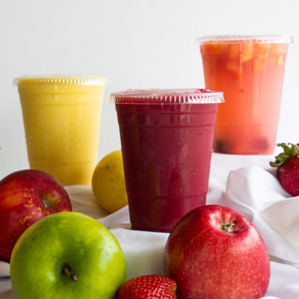 Three colorful Lincoln Grill Cafe smoothies in plastic cups, surrounded by fresh apples, lemon, and strawberries.
